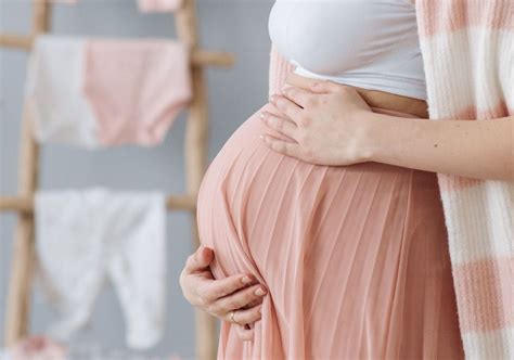 Imagine cu o mamă însărcinată care își ține mâinile pe abdomen, zâmbind.