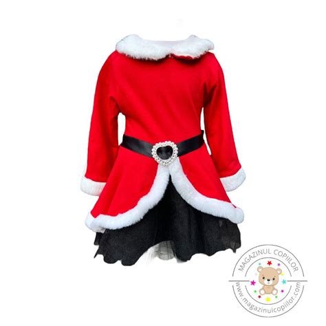 Fetiță îmbrăcată într-o rochiță de Crăciuniță, zâmbind în fața bradului împodobit.