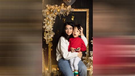 portretul unei mame tinere zâmbind, ținând un copil în brațe, cu un laptop pe masă în fundal